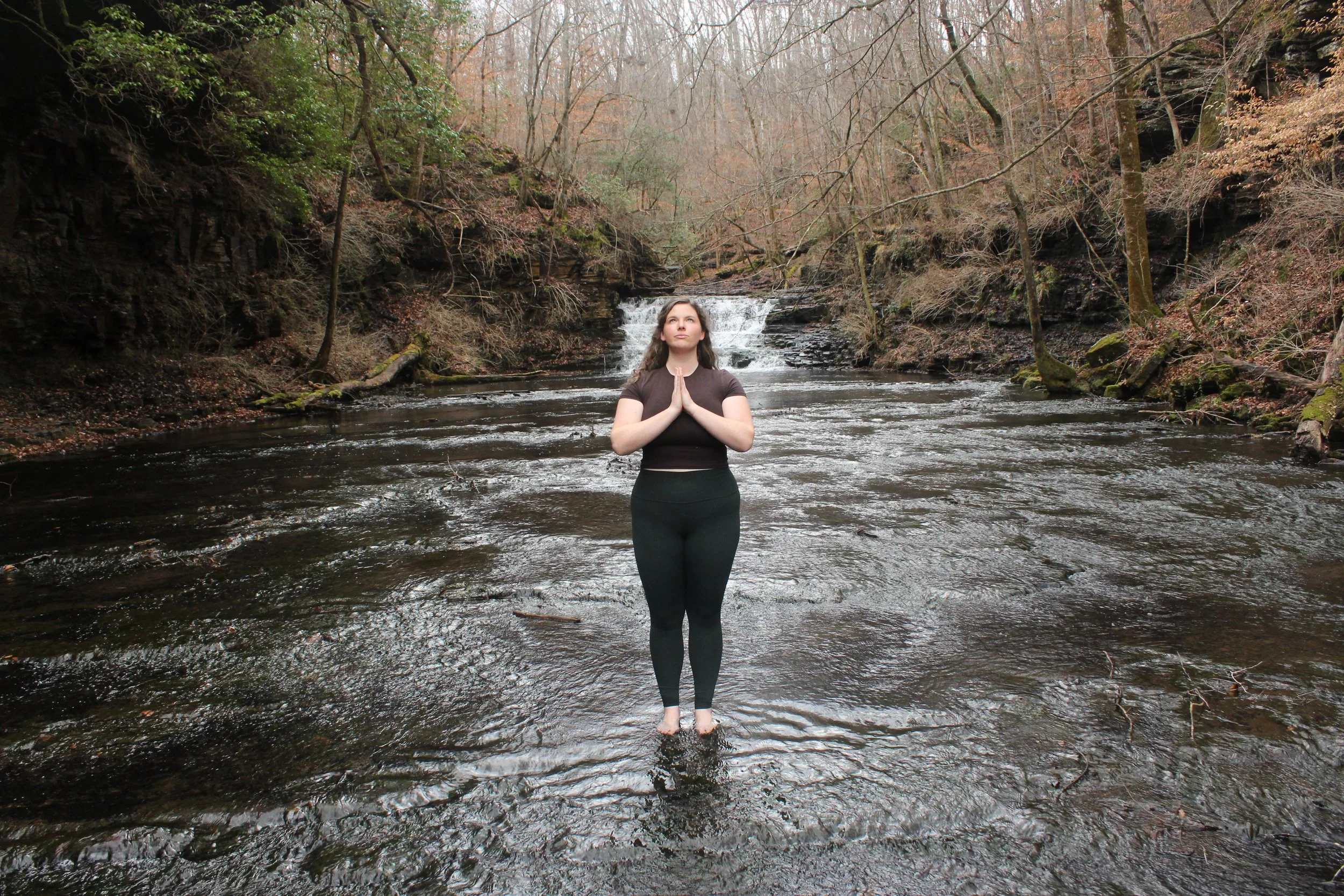 A woman standing in a river with her hands in prayer position, surrounded by trees and a small waterfall in the background.