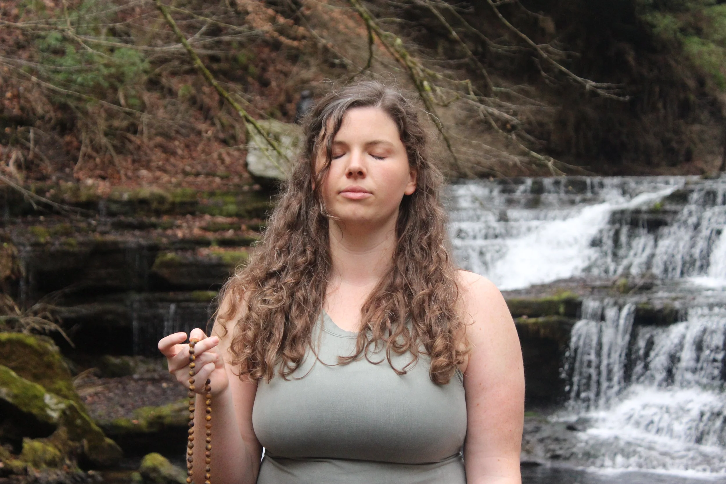 A woman meditating outdoors near a waterfall with her eyes closed, holding prayer beads in her hand.