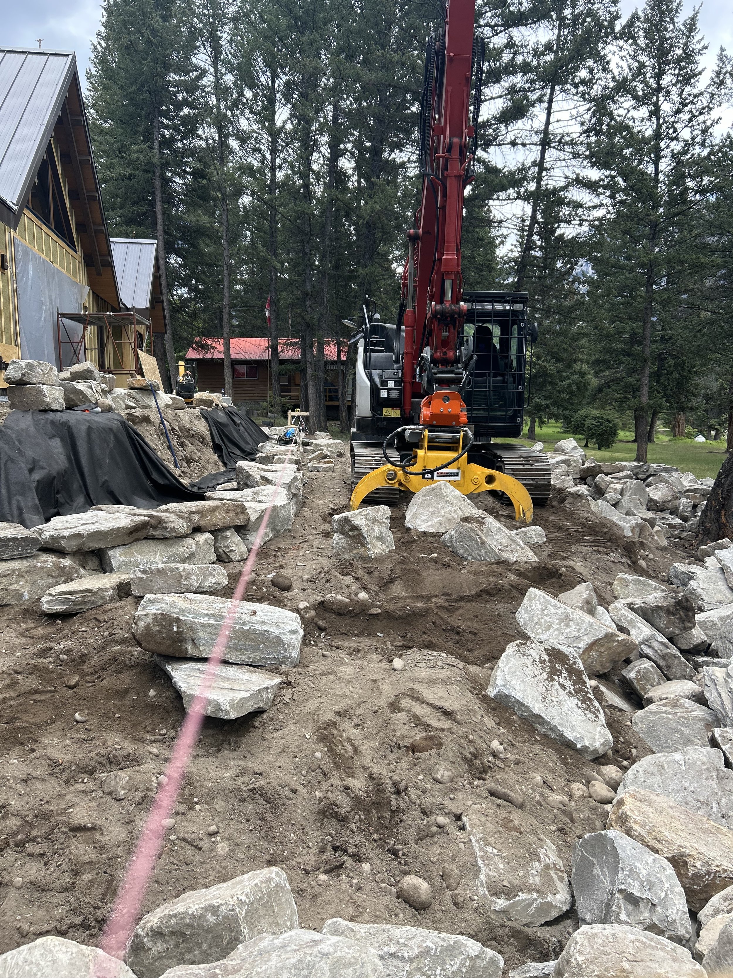 Construction site with a large yellow and black tracked excavator moving rocks, building a stone structure surrounded by trees and a house with a metal roof in the background.