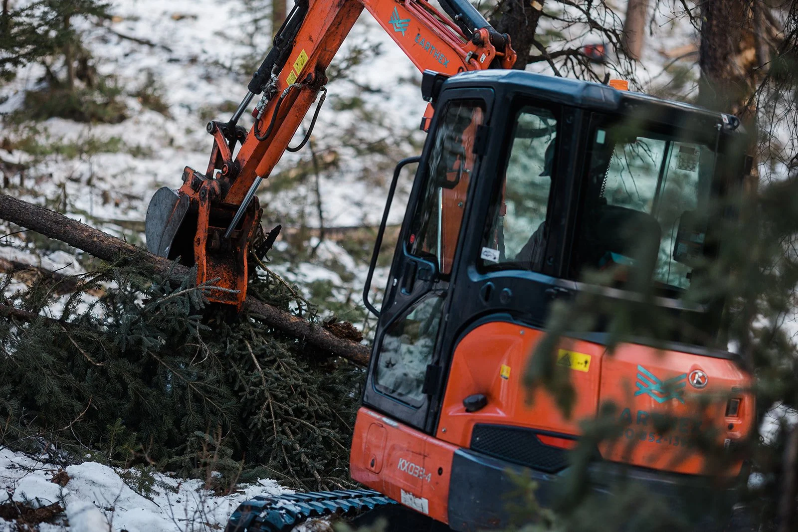 An orange and black excavator cutting a fallen tree in a snowy forest.