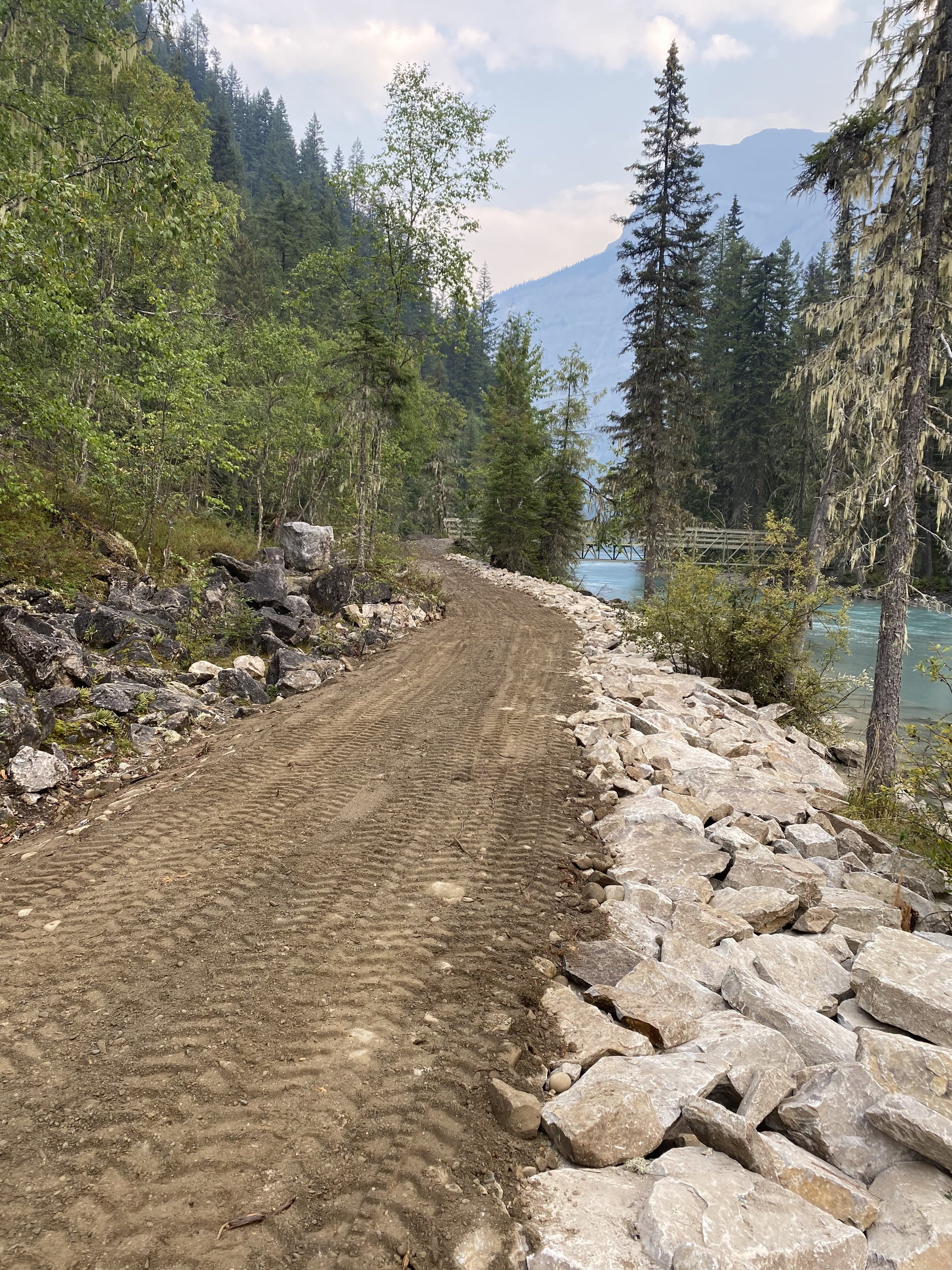 A dirt trail along a river, surrounded by green forest and mountains, with a small bridge in the distance.