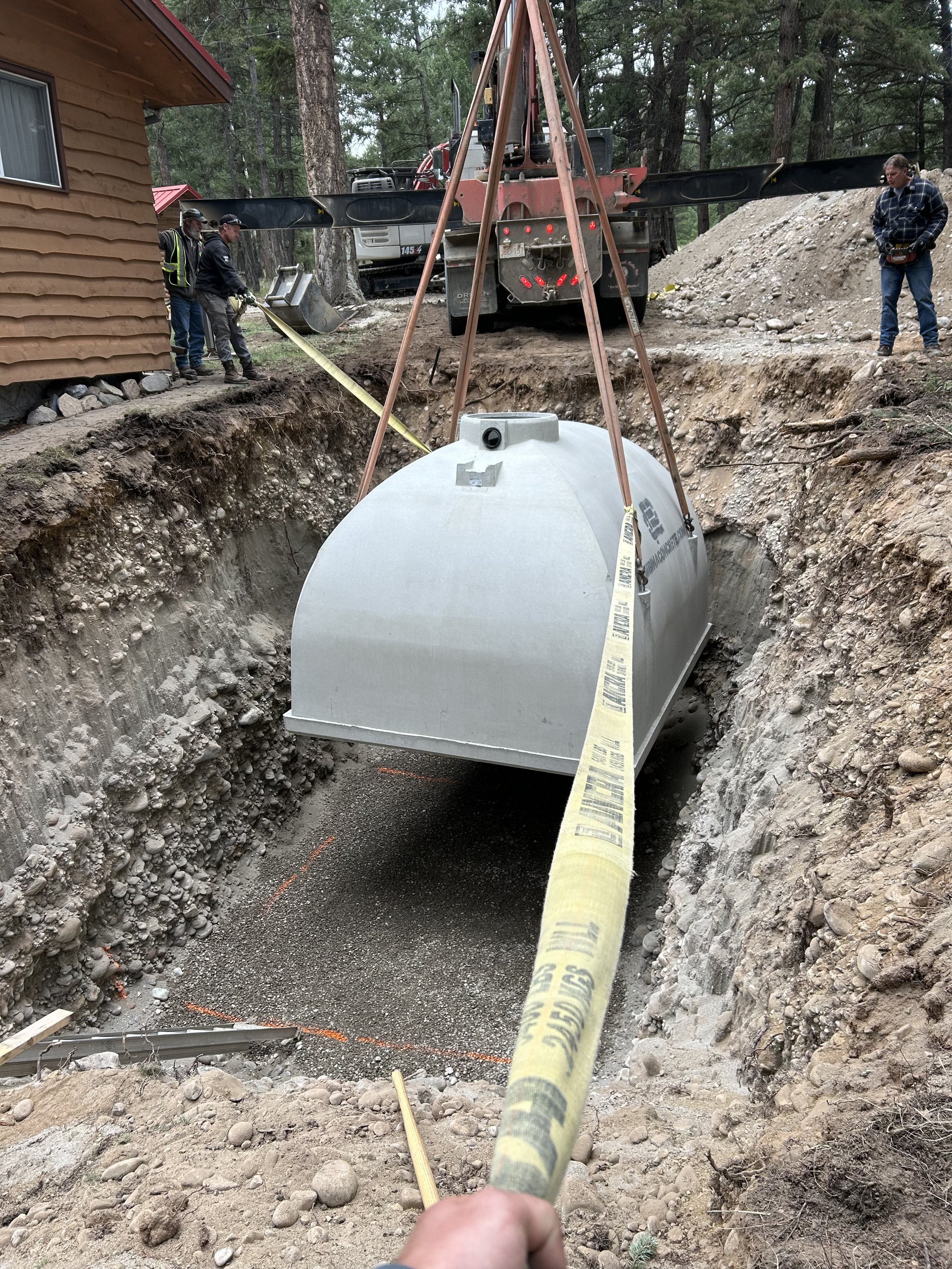 Workers installing a large metal sewer tank into a deep excavated trench with a crane in a wooded area.