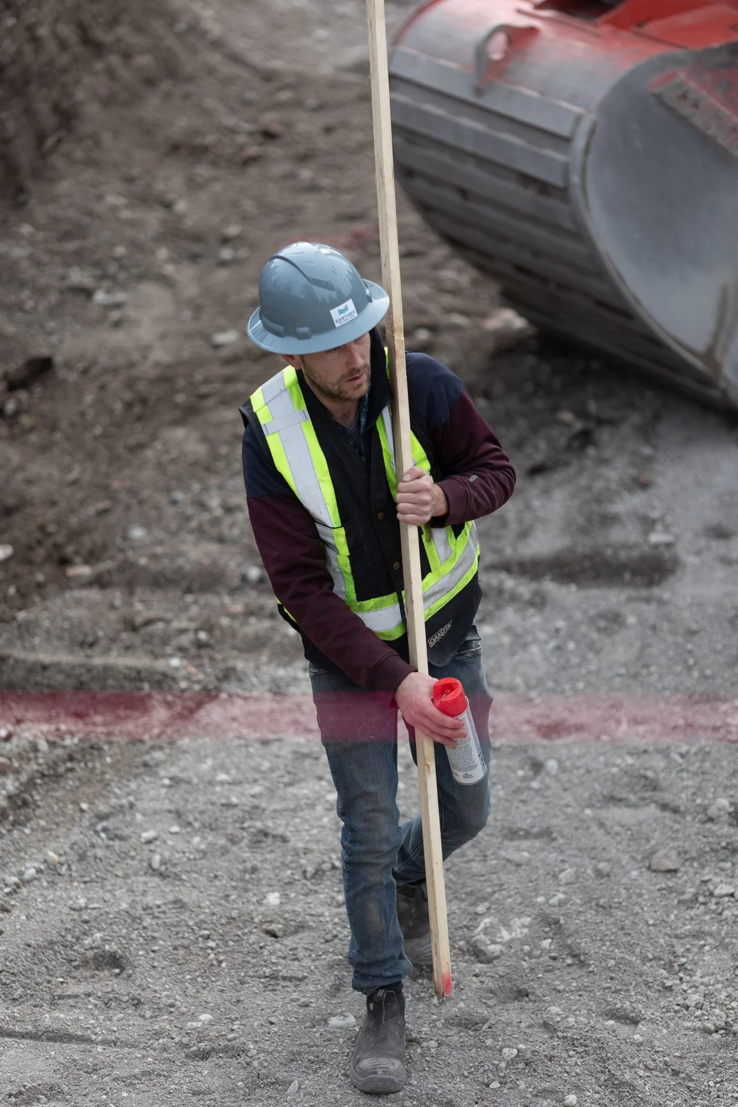 Construction worker wearing a safety helmet, reflective vest, and work boots, holding a long wooden stick and spray can, standing on dirt ground at construction site with machinery in background.