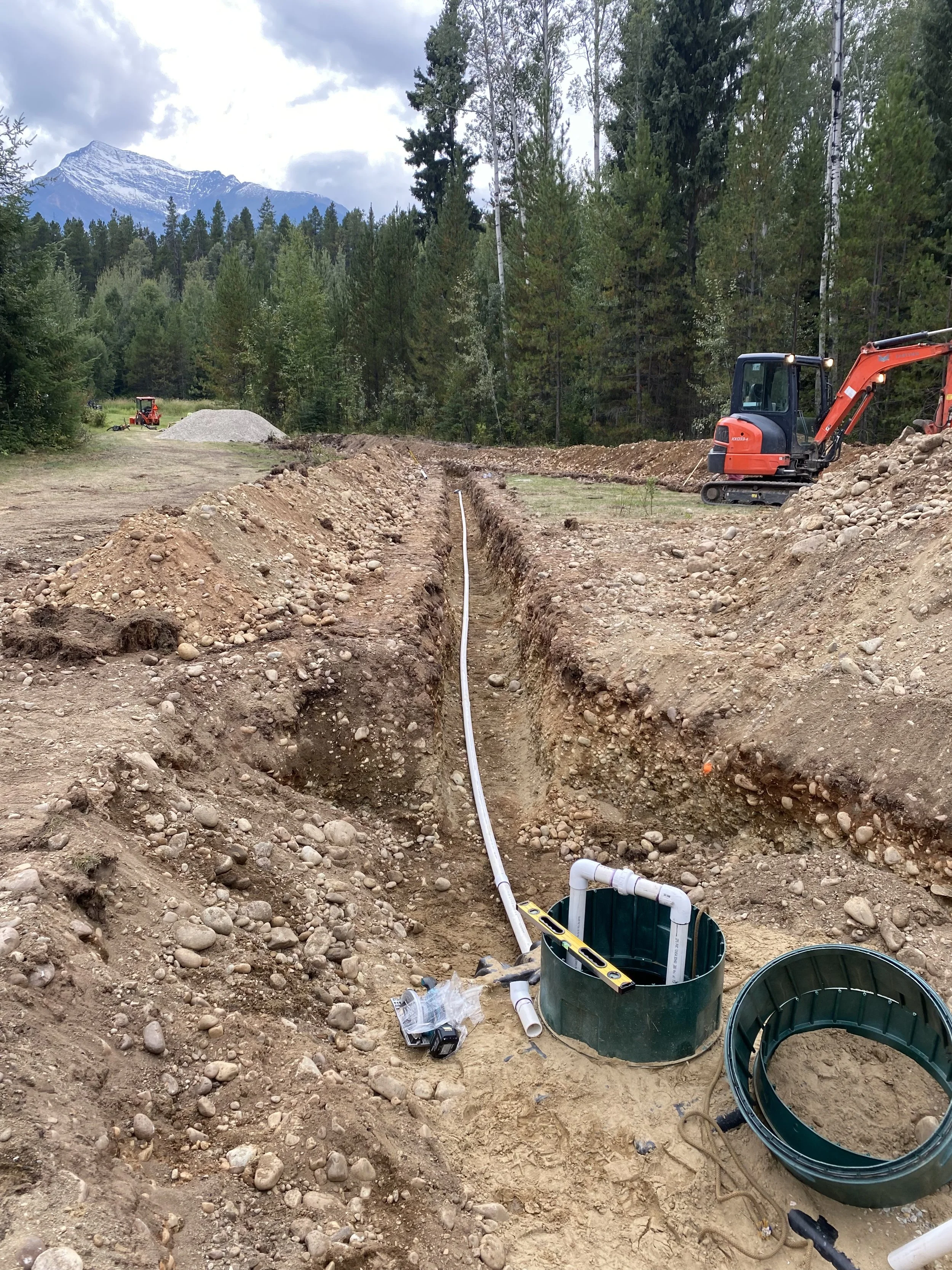 Excavation site with a trench for underground piping, construction tools, and small construction equipment in a natural outdoor setting with trees and mountains in the background.