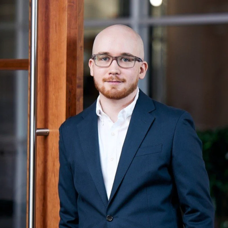 A man with glasses, a beard, and a shaved head, dressed in a navy blue suit and white shirt, standing indoors near a wooden door.