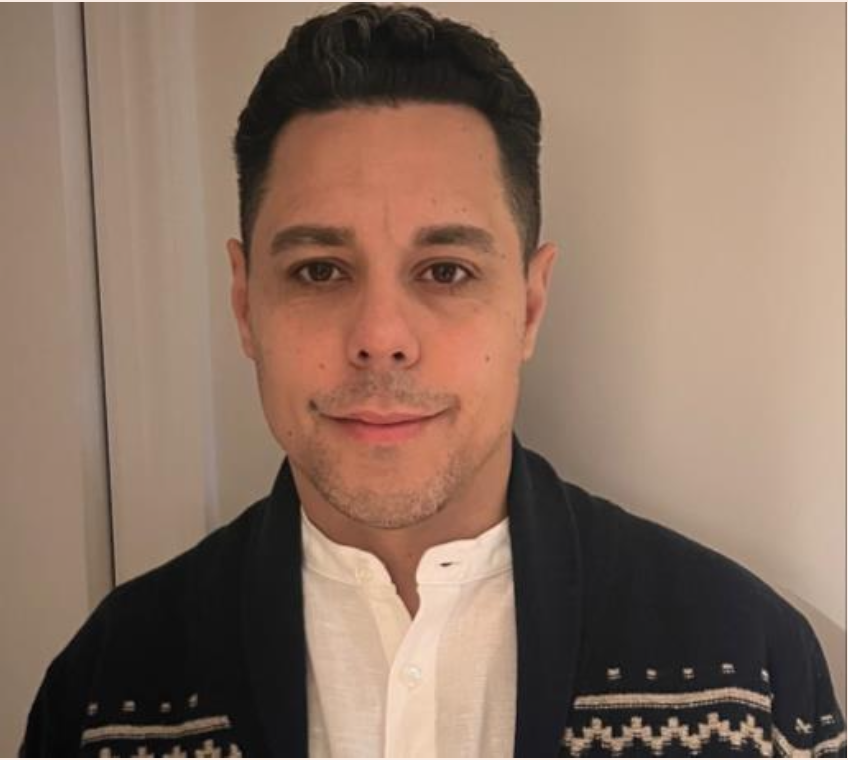 A man with short dark hair and light skin, wearing a white shirt and a black sweater with a patterned design, standing indoors against a plain background, smiling slightly.