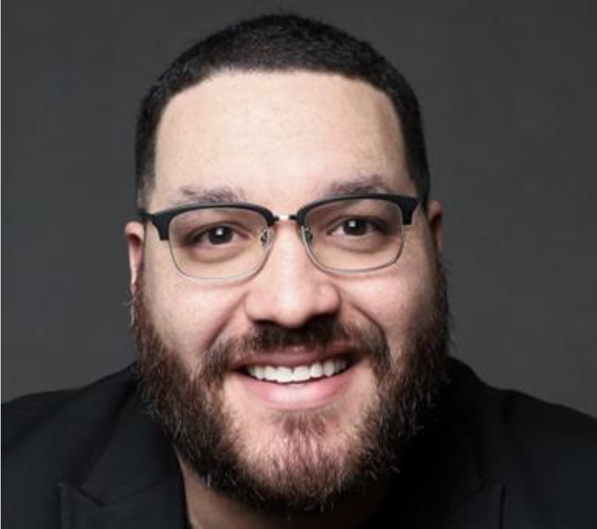 Close-up of a smiling man with glasses, short dark hair, and a beard, wearing a dark suit with a gray background.