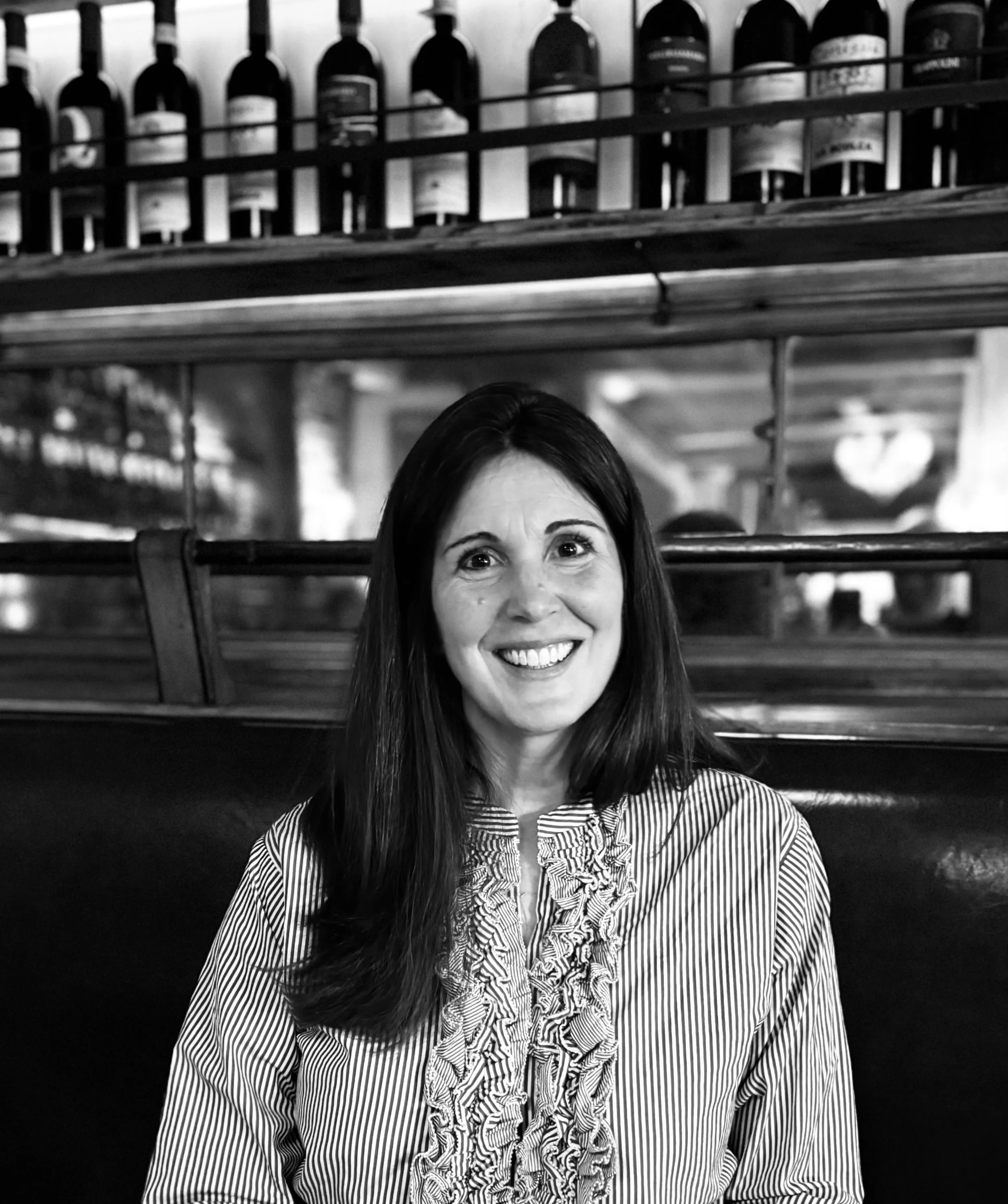 A woman with long dark hair, smiling, sitting in a restaurant or bar with bottles on a shelf above her in the background.