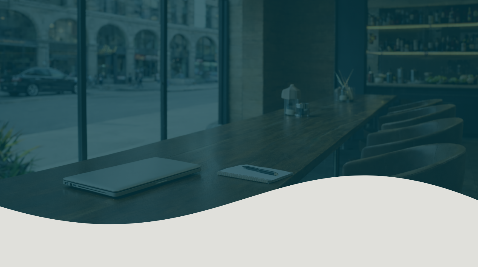 Empty cafe with a long wooden table, laptop, notebook, pen, and chairs near large window showing street view