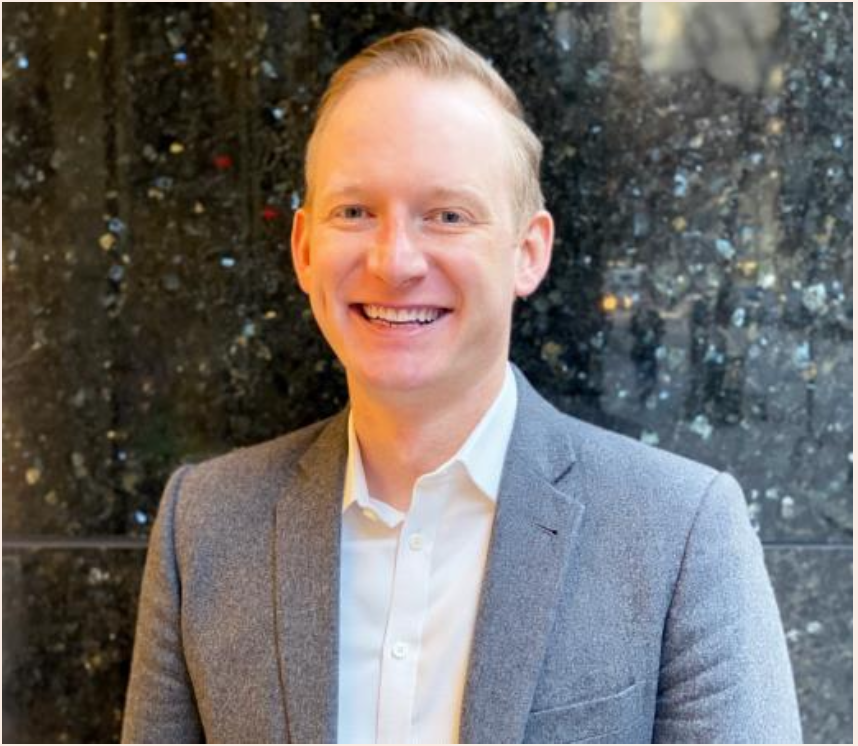 Headshot of a smiling man with blond hair, wearing a gray blazer and white shirt, standing in front of a dark, illuminated background.