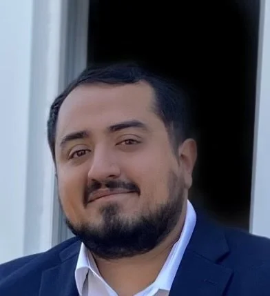A man with dark hair and a beard wearing a suit and white shirt, smiling outdoors in front of a window.