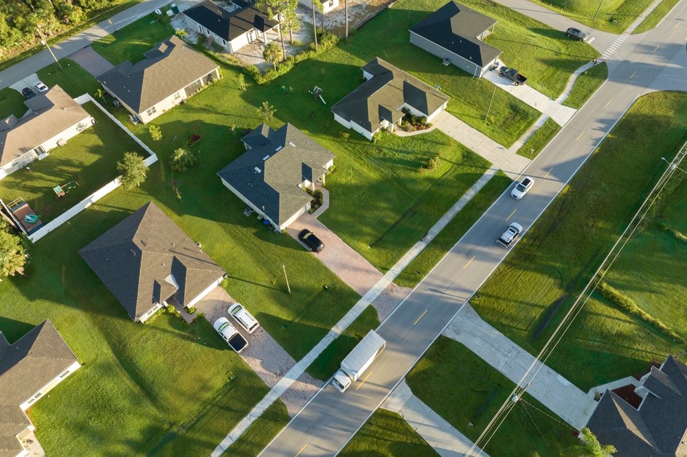 Aerial view of a suburban neighborhood with several houses, green lawns, cars parked on driveways and streets, and power lines.