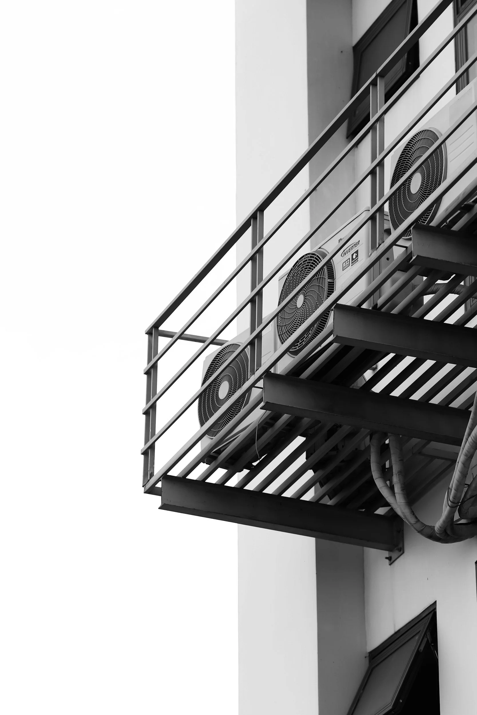 Black and white photo of an exterior building balcony with air conditioning units mounted on the railing and a window below.