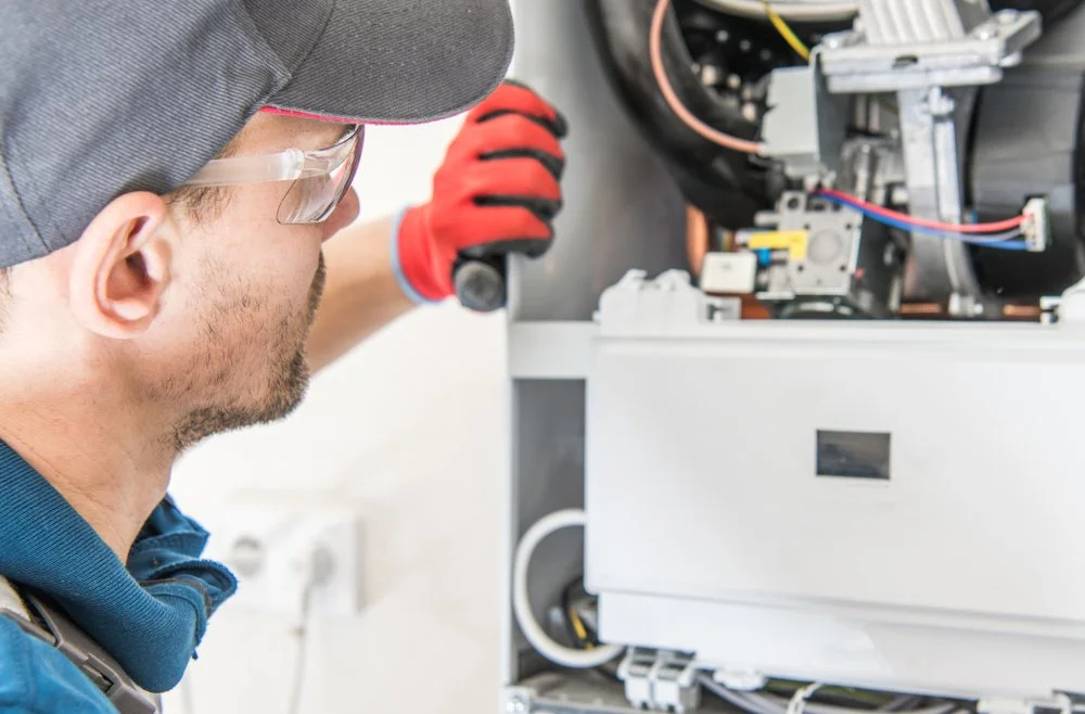 A technician wearing safety glasses, a cap, and gloves inspecting the internal components of a machine or appliance.