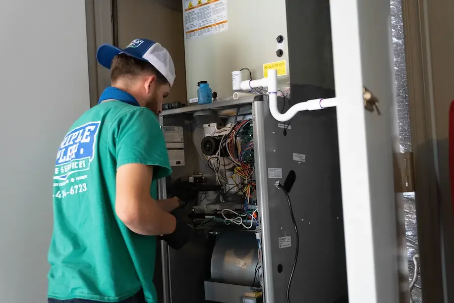 A technician wearing a green t-shirt with company logo and cap working inside an air conditioning or furnace unit, performing maintenance or repair.