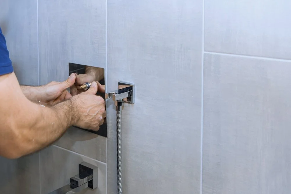 A person installing a chrome shower faucet on a light gray tiled wall.