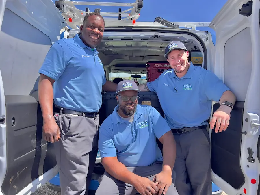 Three men in blue polo shirts and hats smiling at the camera inside the back of a white van with a ladder on top, outdoors on a sunny day.