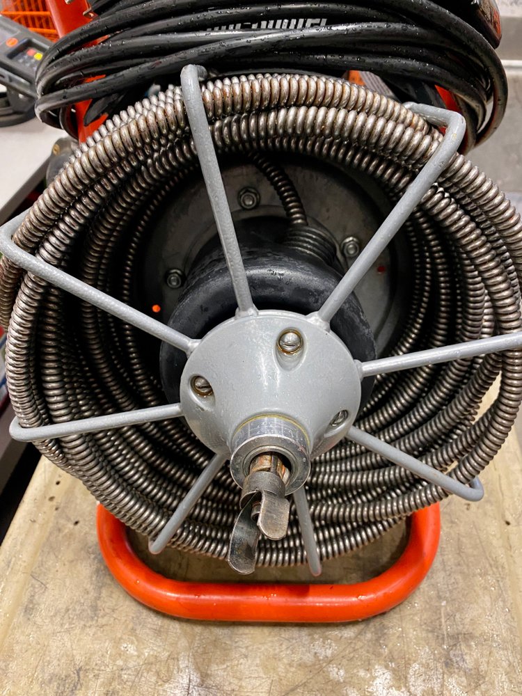 A close-up view of a coiled industrial hose reel mounted on a workbench with metal supports, surrounded by black cables and orange handles.