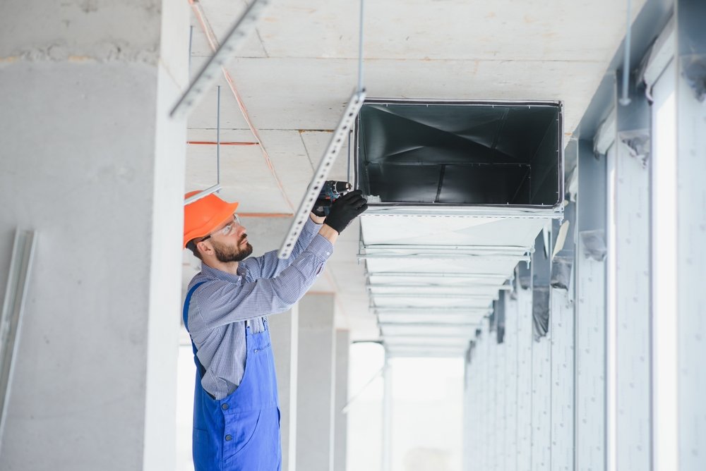 A construction worker wearing an orange helmet, blue overalls, and glasses, working on installing or inspecting an HVAC duct in a building under construction.
