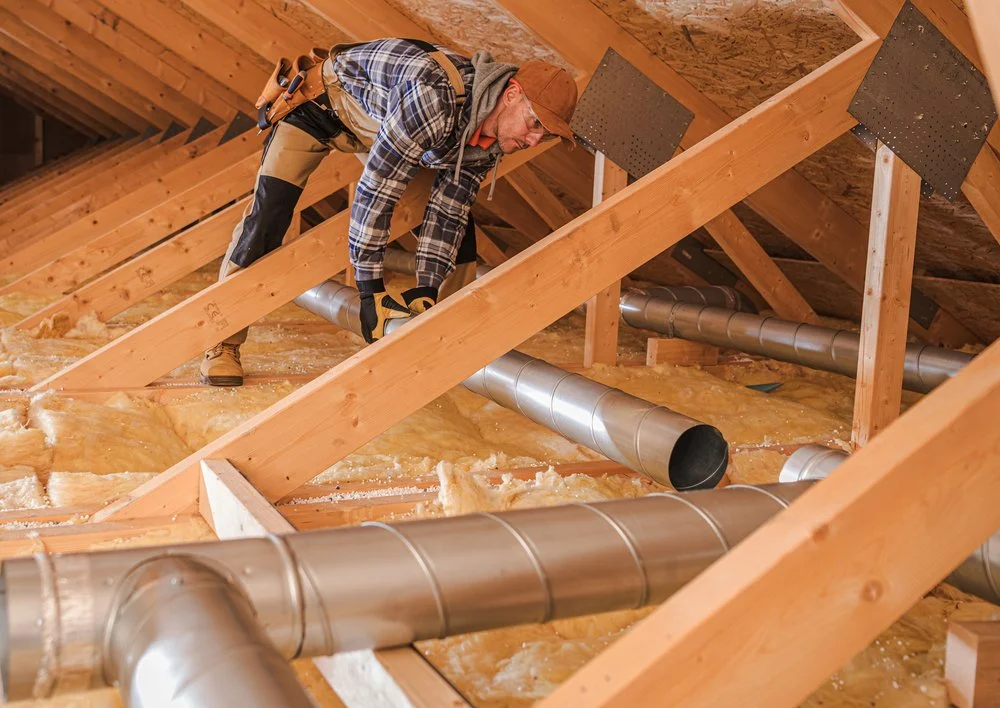 A man wearing a plaid shirt, jeans, and a hat is working on installing ducts in an attic space framed with wooden beams, insulation, and ventilation pipes.