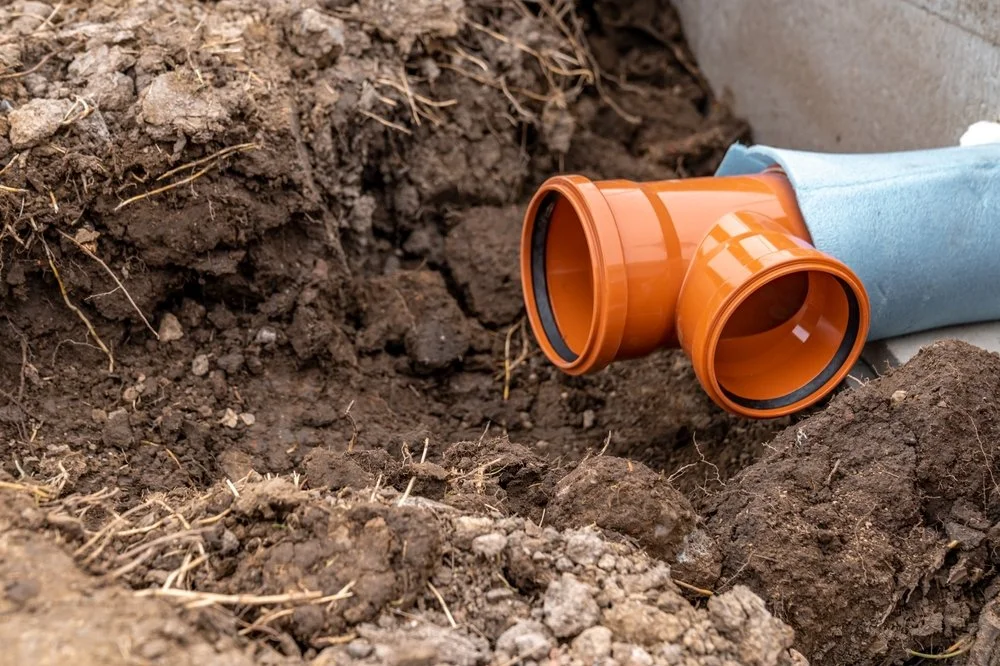 Close-up of a dug trench with orange PVC pipe fittings and a piece of blue pipe, with dirt and soil surrounding them, possibly for plumbing installation.