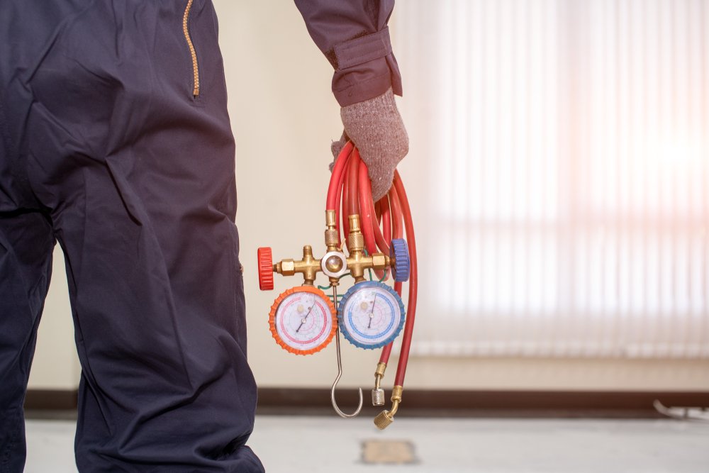 Person wearing work gloves holding a manifold with red and blue hoses, gauges, and a valve.