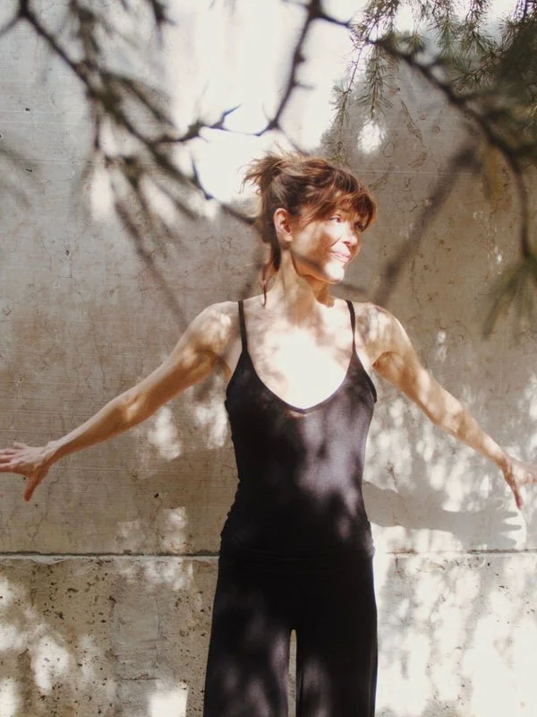 A woman standing outdoors against a concrete wall with her arms outstretched, smiling, and surrounded by tree shadows and branches.