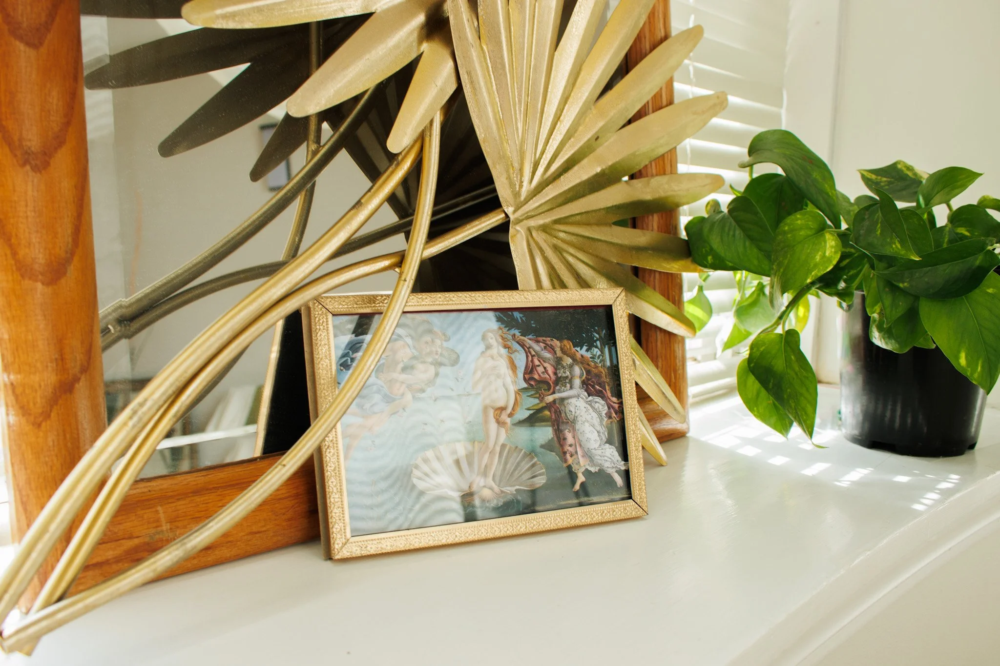 Decorative mirror, gold and black paper fans, framed religious artwork, and potted green plant on white shelf near window with blinds.