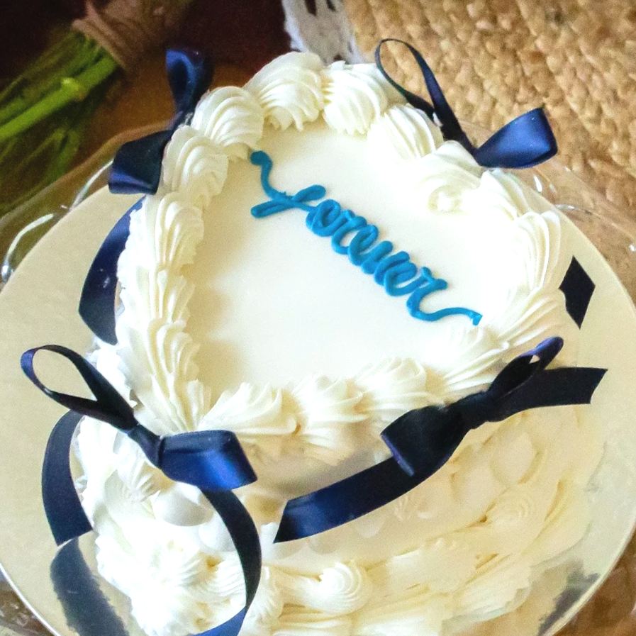 A heart-shaped white frosted cake with the word 'LOVE' written in blue on top, decorated with black ribbons on the edges.