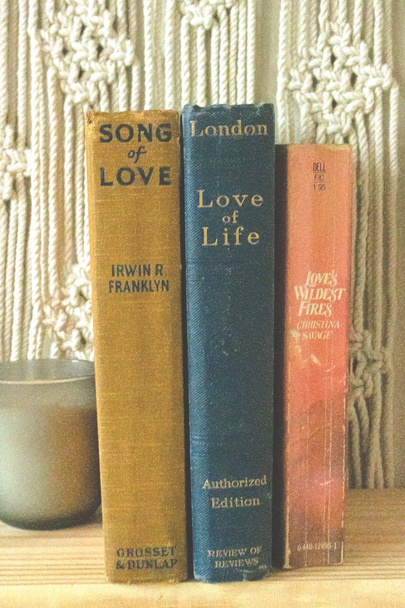 Three books on a wooden shelf, with a gray ceramic pot to the left. The books are titled "Song of Love" by Irwin R. Franklyn, "London Love of Life" by unknown author, and "Love's Wildest Fires" by Christina S. S. King.