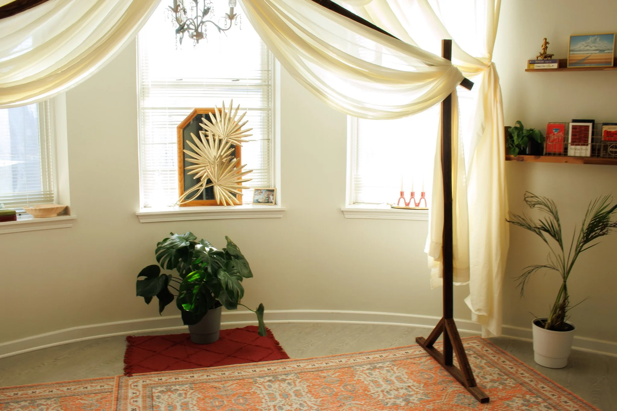 A bright living room with cream-colored walls and large windows with white blinds. There are cream-colored curtains draped over the windows. A chandelier hangs from the ceiling. On the windowsill, there's a framed photo and a decorative wooden palm l