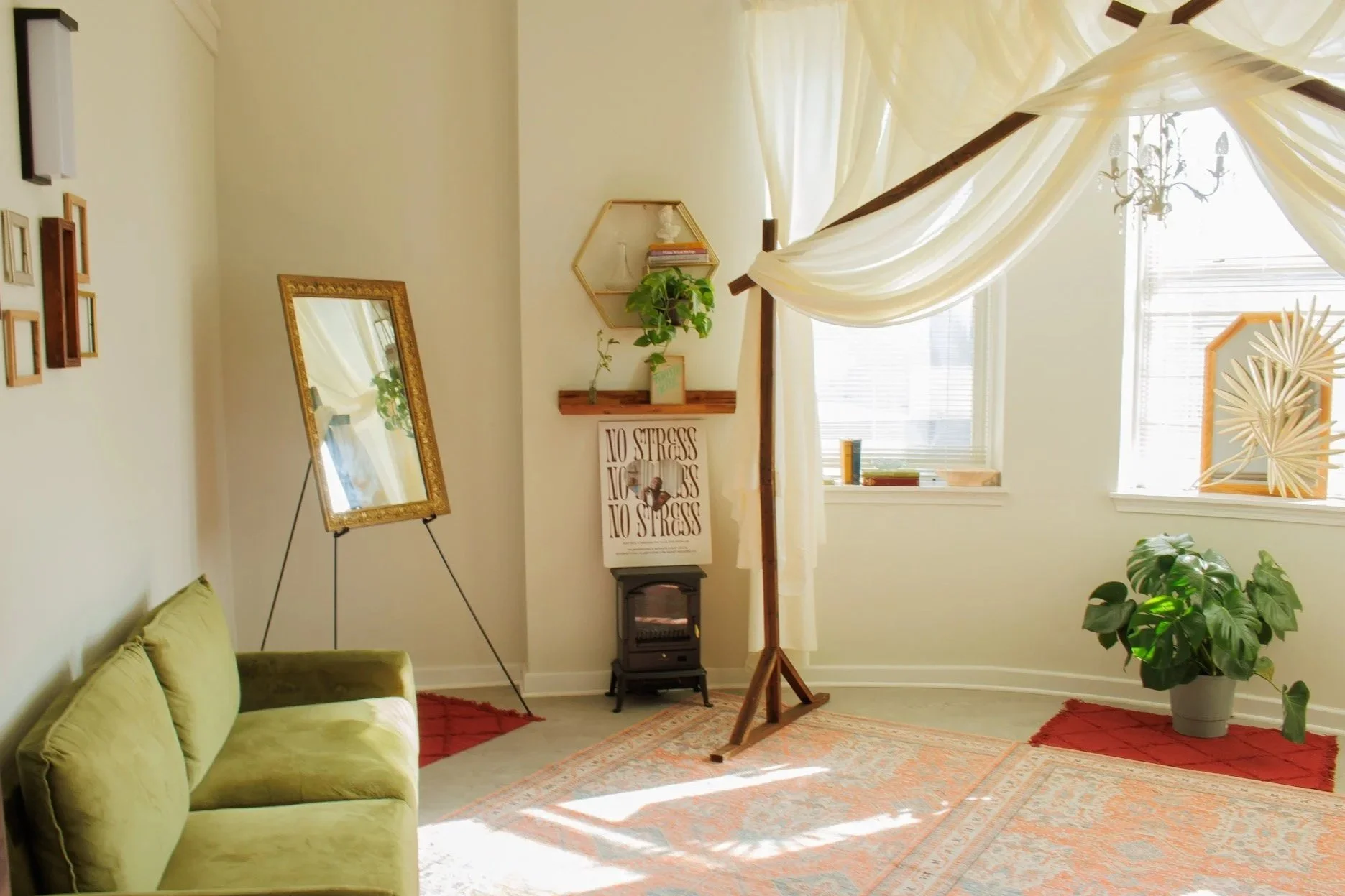 Bright, cozy living room with a green velvet sofa, a large mirror on a stand, a small wood stove, potted plants, and decorative wall art, illuminated by natural light through two large windows covered with sheer curtains.