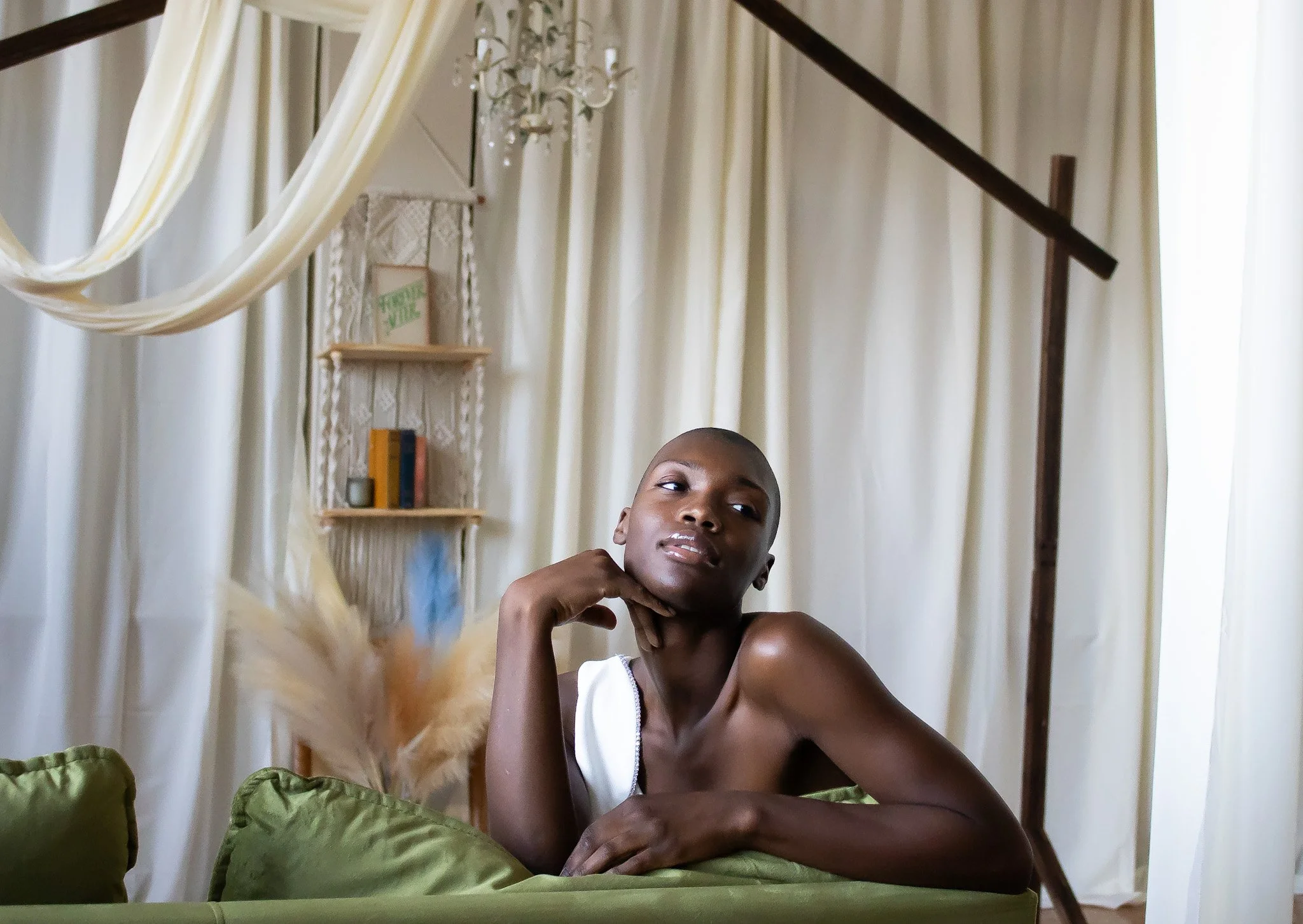 A person with a shaved head sitting on a bed with green pillows, in a room with cream-colored curtains, a chandelier, and shelving behind them.