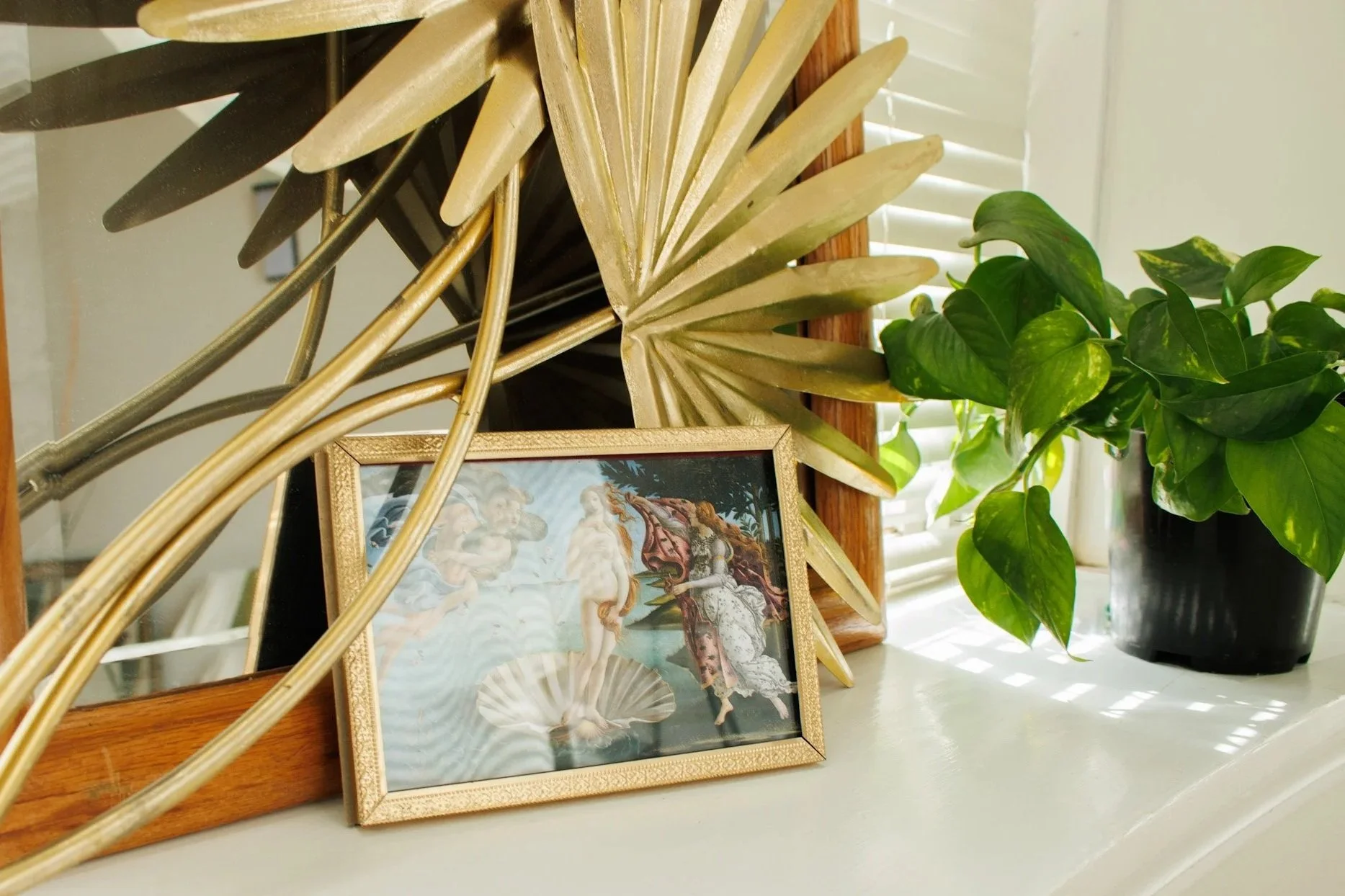 Decorative gold framed picture of a woman and cherubs, a large gold palm leaf, and a green potted plant on a white shelf in front of a window with blinds.