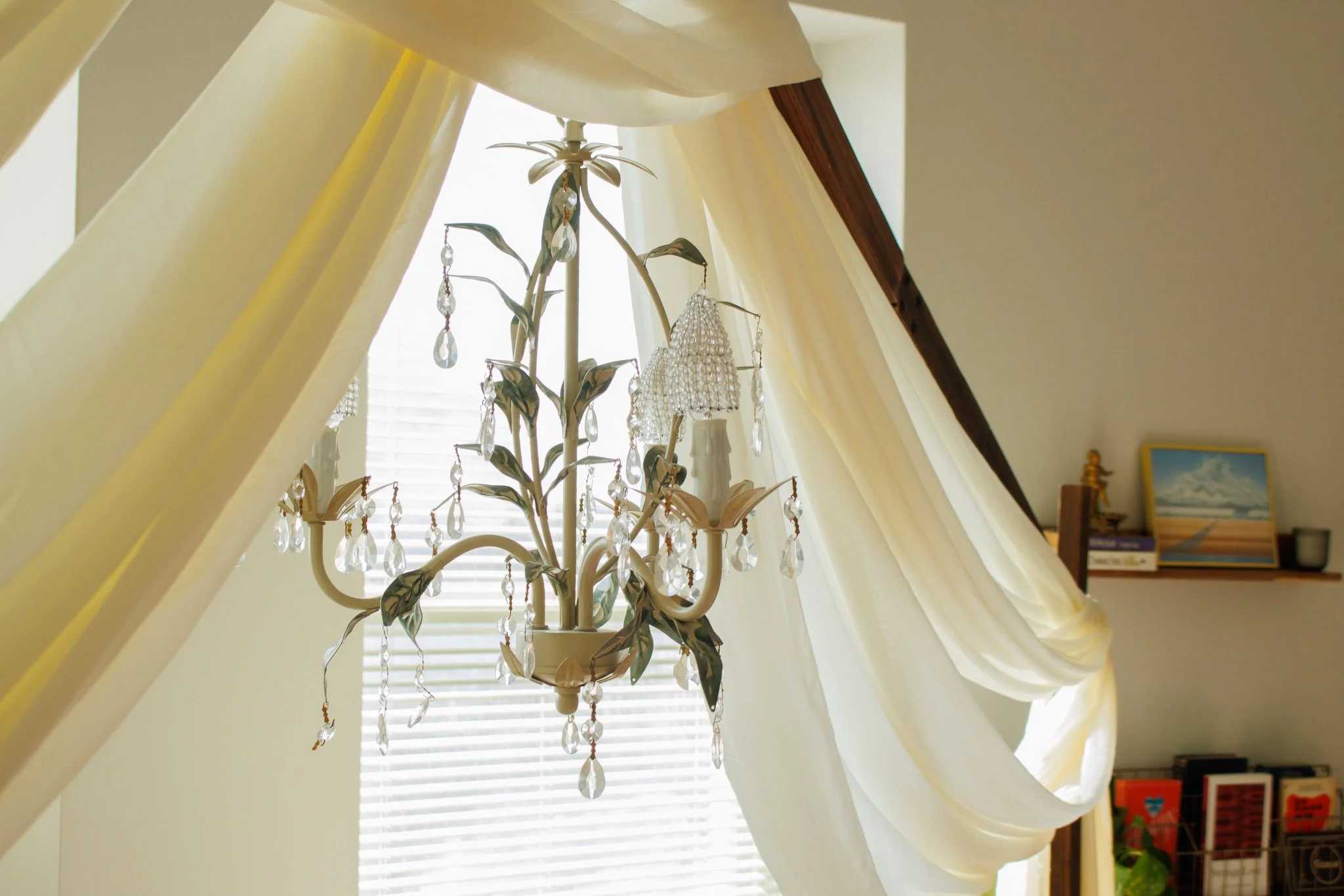 A chandelier with crystal ornaments and decorative metal branches hanging in a room with cream curtains and blinds, with a shelf holding books and framed pictures in the background.