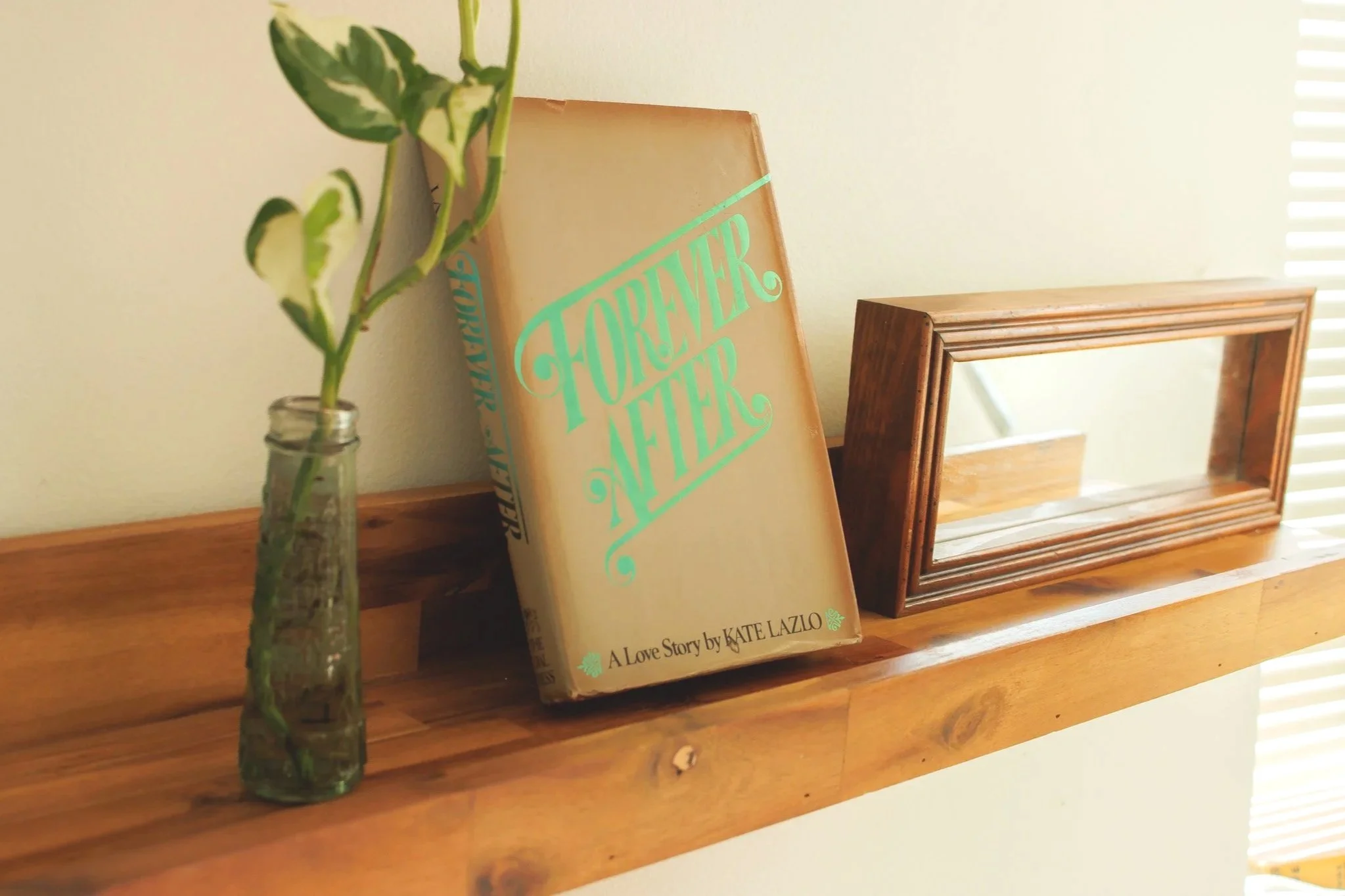 A wooden shelf with a clear glass vase holding a leafy plant, a book titled 'Forever After' by Kate Lazio, and a wooden framed mirror, with blinds and sunlight in the background.
