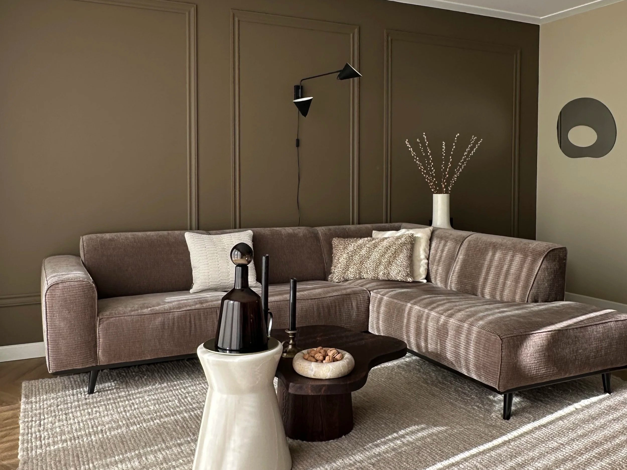 Living room with a large beige sectional sofa, decorative pillows, a round dark wood coffee table with nuts, a modern white vase with branches, a wall-mounted black lamp, and beige walls with decorative panels and art.