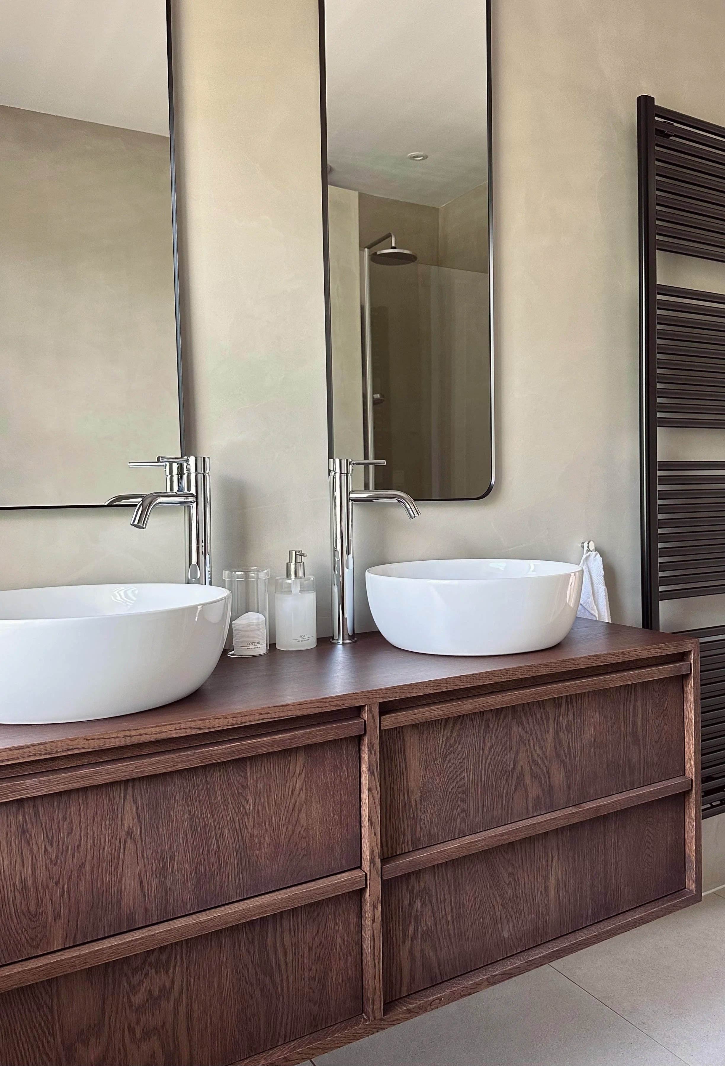 Modern bathroom with a double wooden vanity, two white vessel sinks, and silver faucets. A large mirror and a shower area with a rainfall showerhead are visible in the background, along with a black heated towel rail on the wall.