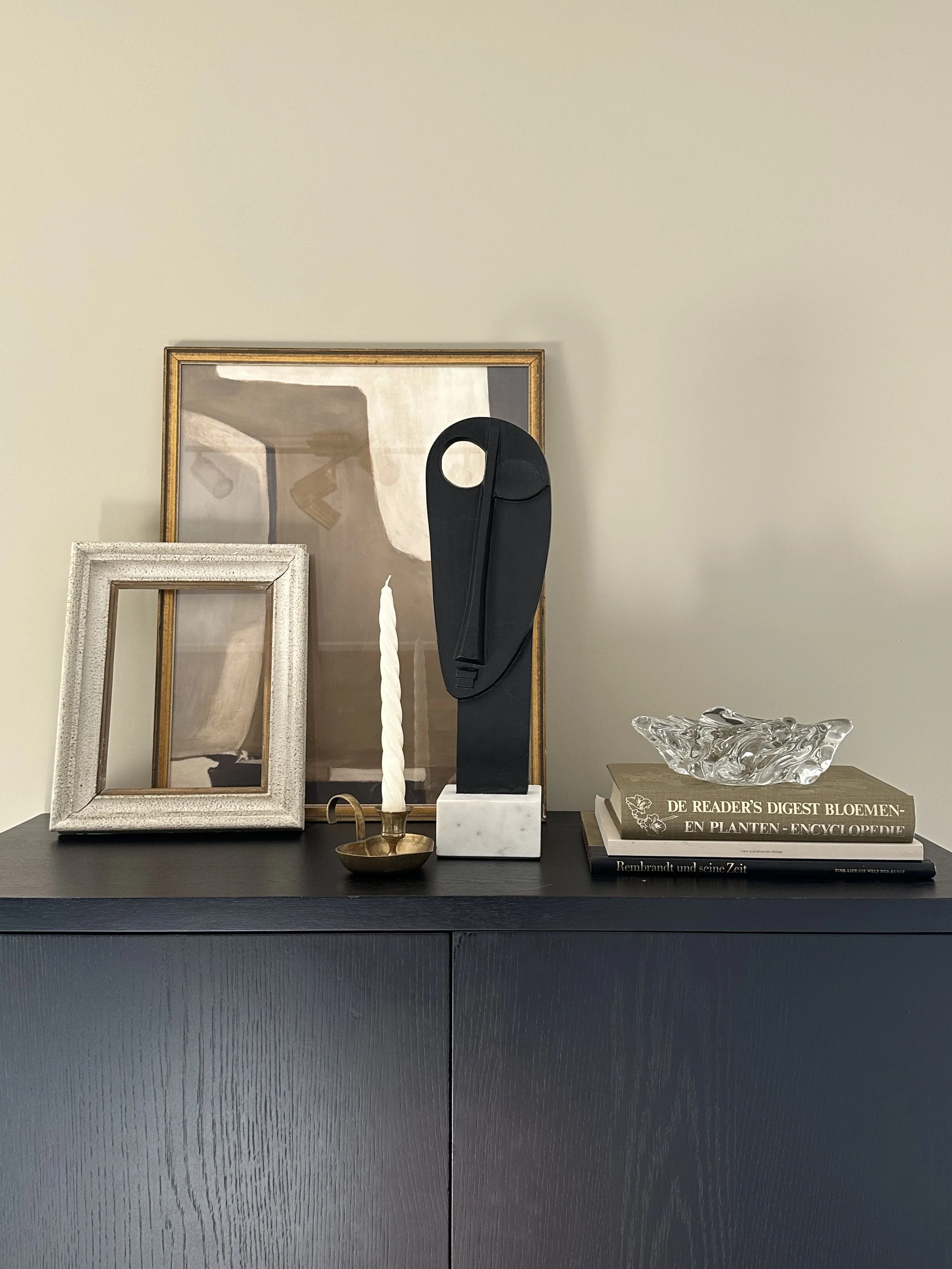 Decorative items on a black cabinet, including a large framed abstract artwork, a small empty silver frame, a twisted white candle in a brass holder, a black abstract sculpture, a glass decorative bowl, and a stack of books.