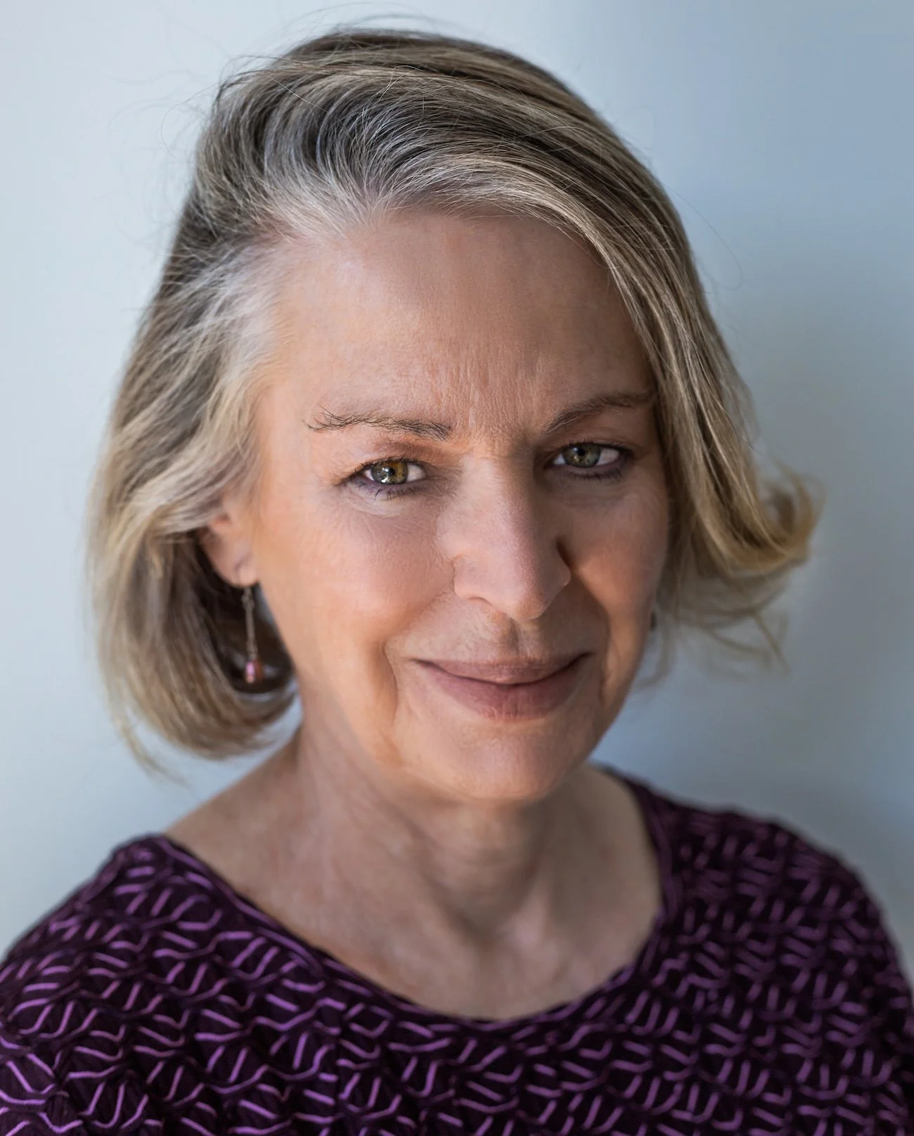 A middle-aged woman with short, wavy gray hair and light green eyes, wearing a purple patterned top and earrings, smiling softly.
