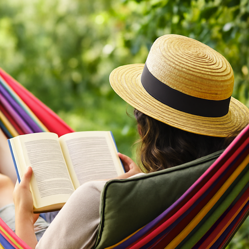 Person wearing a straw hat reading a book while relaxing in a hammock outdoors.
