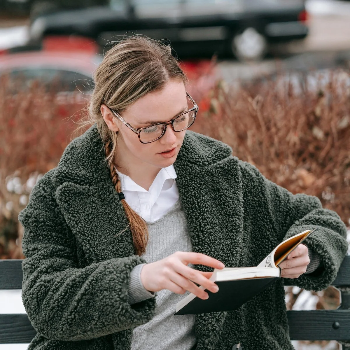 A young woman with glasses and braided hair, wearing a dark fuzzy coat, reading a book while sitting on a bench outdoors during fall.