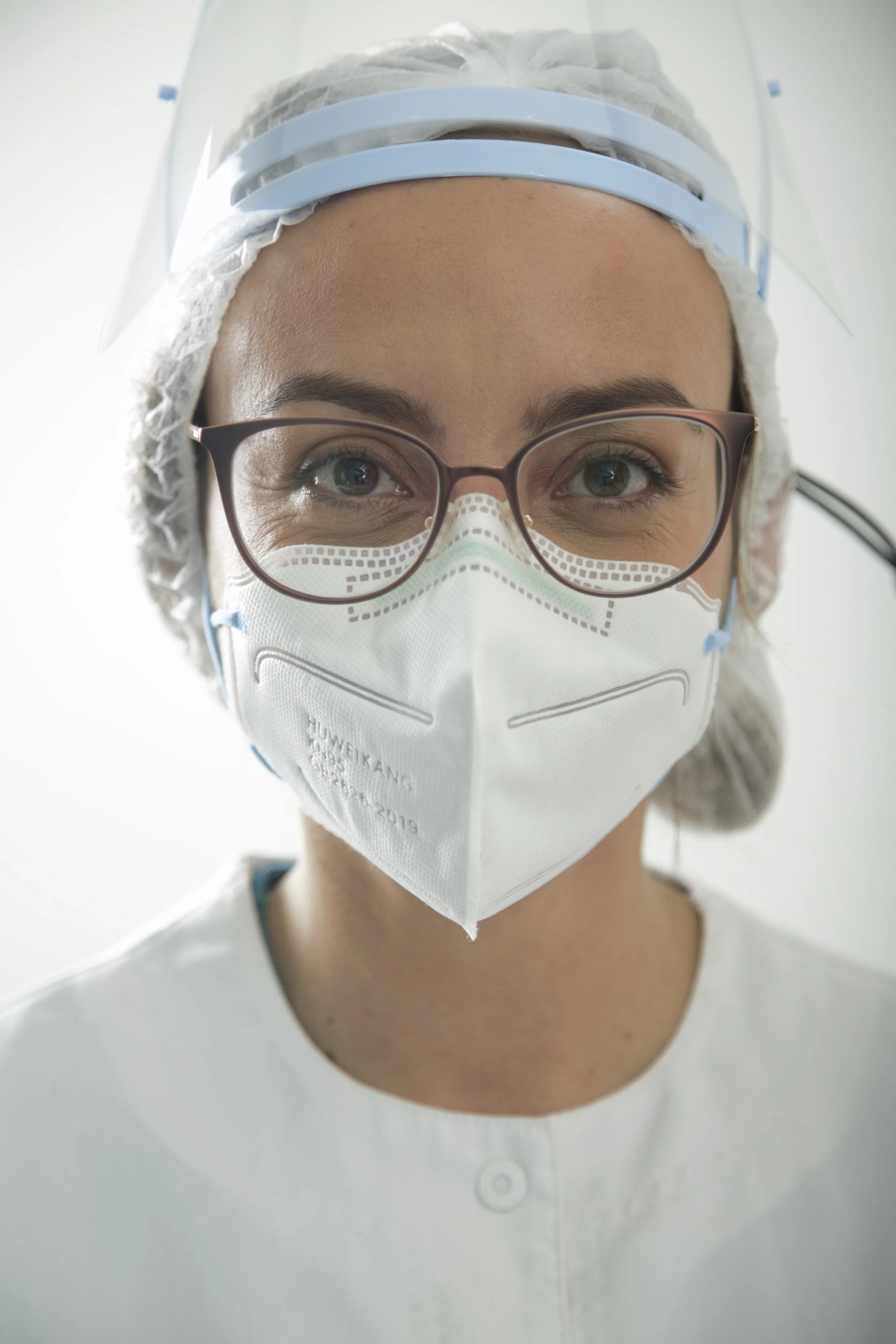 Médico con gafas, mascarilla facial y protección de cabeza, mirada directa a la cámara.