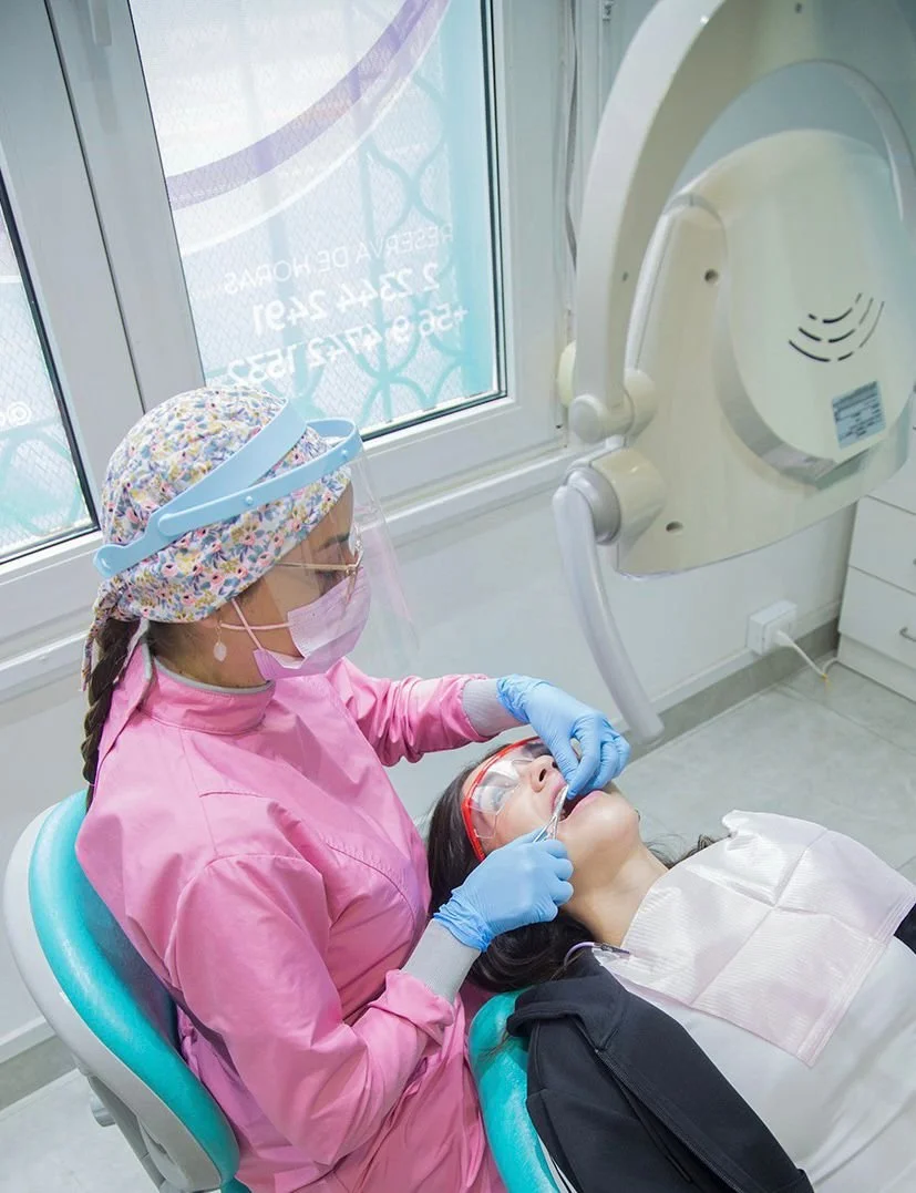 Una dentista atendiendo a una paciente en un consultorio dental, con equipo de protección y radiografía en una máquina de rayos X.