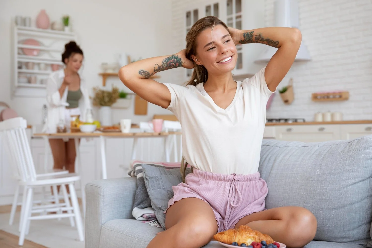 Mujer sonriendo con las manos en la cabeza sentada en un sofá en una sala moderna, con una mesa con desayuno frente a ella y otra mujer en la cocina en el fondo.