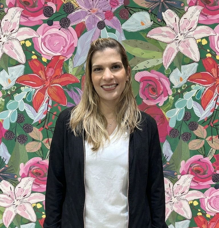 Una mujer sonriente con cabello rizado y oscuro, vestida con una blusa negra, de fondo un mural con flores de diversos colores, incluyendo rosas, lirios y otras plantas.