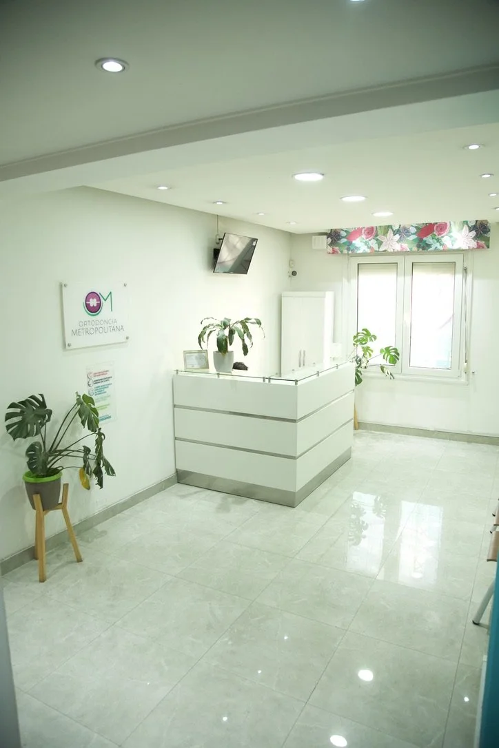 Recepción de una clínica de ortodoncia con plantas decorativas, una ventana con cortina floral, y un cartel que dice 'Ortodoncia Metropolitana'.