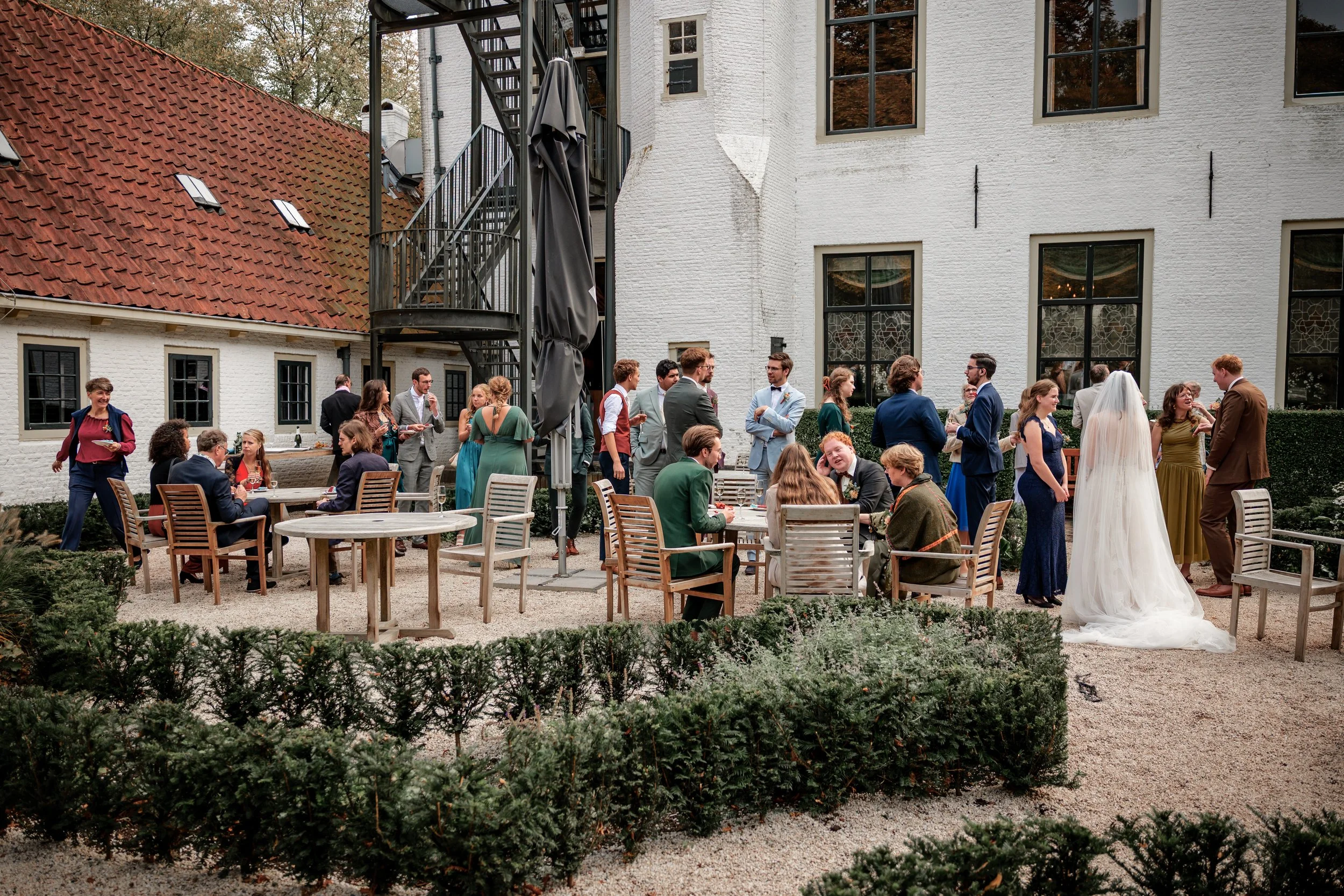 Een groep mensen die op een binnenplaats samen is voor een bruiloft. Er zitten mensen aan tafels en anderen staan in kleine groepen. Een bruid in witte trouwjurk staat bij een man in een bruin pak. Gefotografeerd door professionele fotograaf.
