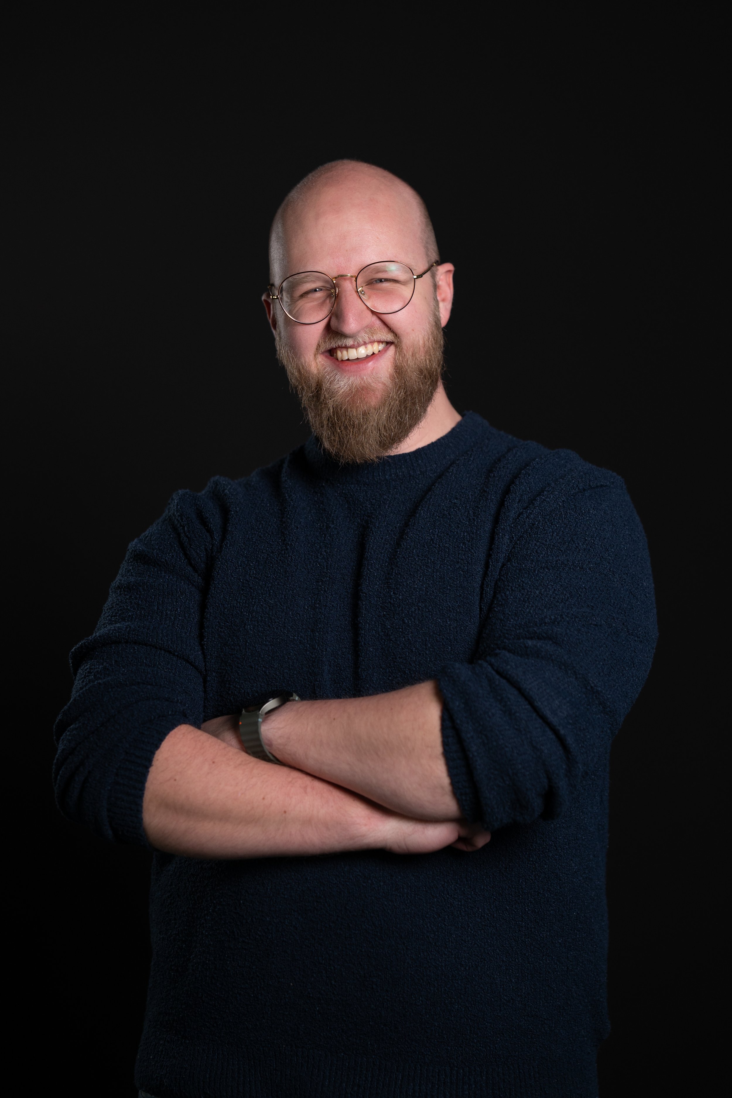 Man met een bril, een baard, en een glimlach, gekleed in een donkerblauwe trui, staand met gekruiste armen tegen een zwarte achtergrond.