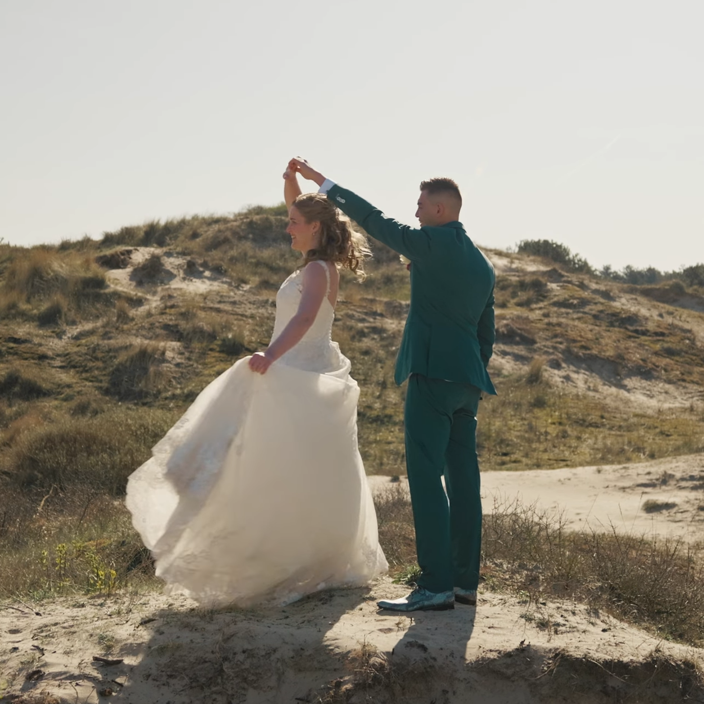 Een stel dat danst op een zandstrand met duinen op de achtergrond. De vrouw draagt een witte trouwjurk en de man een blauwe pak. Shot uit cinematic trouwfilm in Nederland